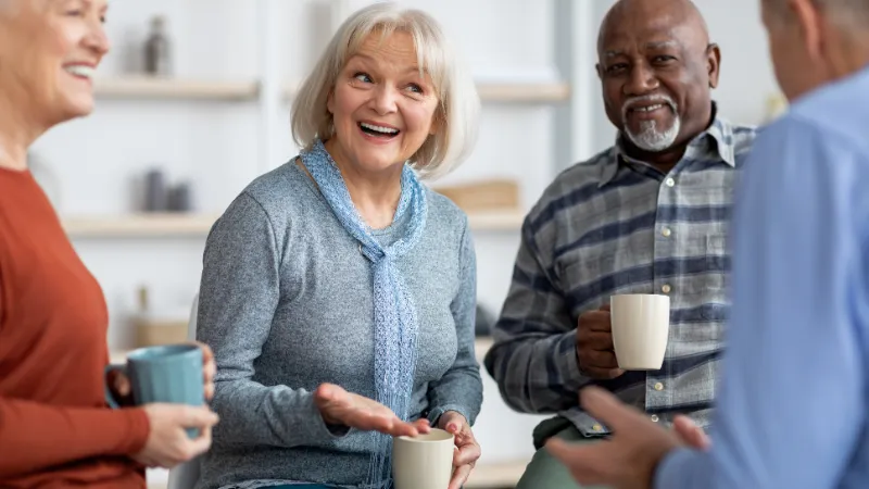 Four people standing together in a community setting, talking and holding hot drinks during a social gathering.